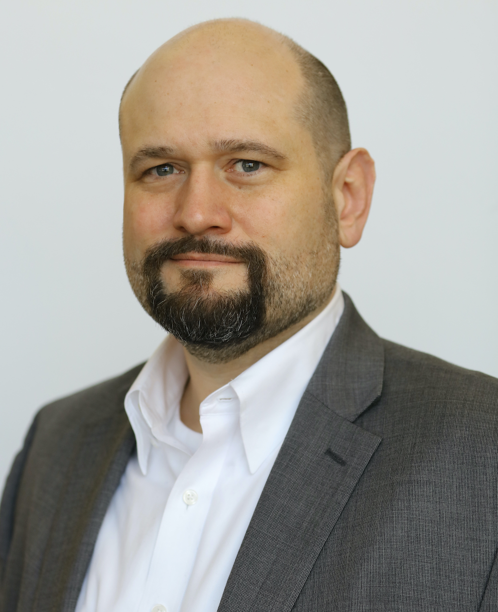 Middle aged man with shaved head and goatee wearing a white Oxford shirt and gray sport coat smiling wryly at the camera.
