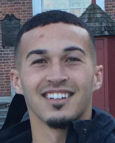 Young man with short cropped hair smiling at the camera in front of a brick building.