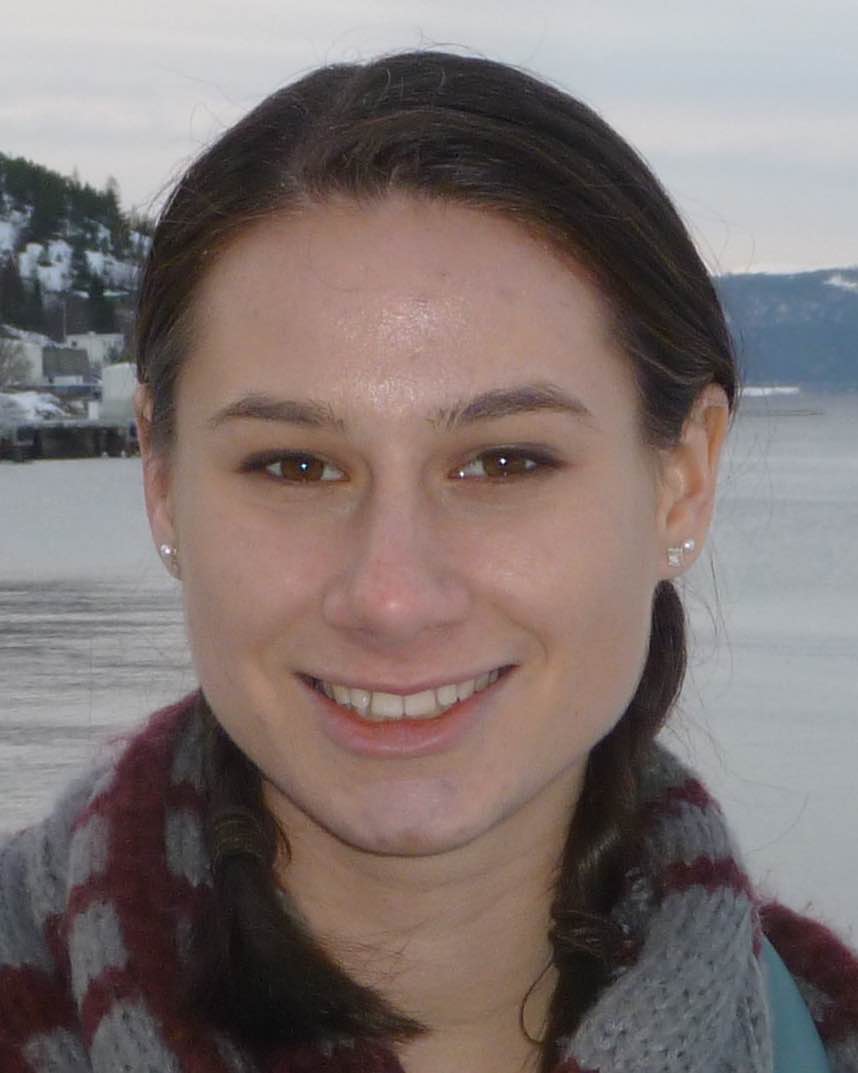 A smiling young woman with dark hair and a gray and burgundy scarf standing in front of a calm lake with snow covered hills and a gray sky in the background.