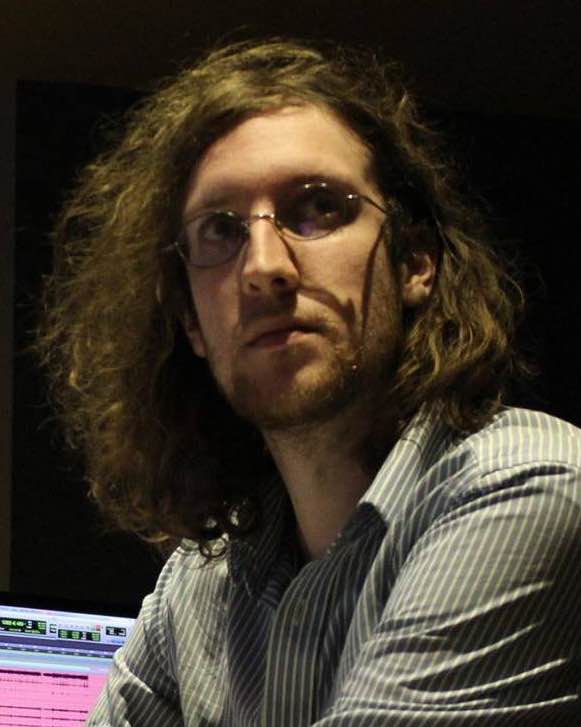 A tall, bespectacled man with long, wavy hair and a light beard looks past the camera out of the corner of his eye.